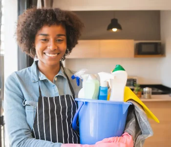 Portrait of young afro woman holding a bucket with cleaning items at home. Housekeeping and cleaning concept.