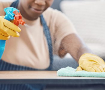 Woman with detergent to clean, spray and wipe table, in office or living room in home. Black woman in domestic work, use cloth and liquid cleaner, on desk for hygiene in lounge of business or house.